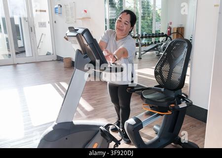 Eine Frau ist in einem Fitnessstudio auf einem Fitnessrad am Trainieren. Der Raum ist hell mit großen Fenstern. Sie wirkt konzentriert auf ihre Fitnessroutine. Stockfoto