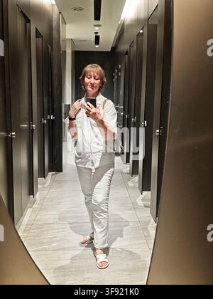 Eine Frau steht vor einem Spiegel, lächelt und hält ein Smartphone. Sie fängt einen Moment in einer Toilette ein, umgeben von Türen. Stockfoto