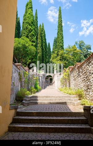 Sonnige steinerne Treppe, die durch einen ummauerten Gartenweg zu Zypressen und einem gewölbten Tor im Il Vittoriale nahe dem Gardasee in Italien führt Stockfoto