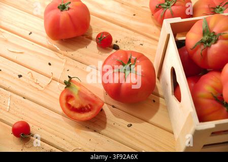 Reife Tomaten ruhen auf einer sonnenbeschienenen Holzoberfläche neben einer rustikalen Kiste, wobei eine geschnittene Tomate ihr saftiges Inneres und die Samen offenbart. Stockfoto
