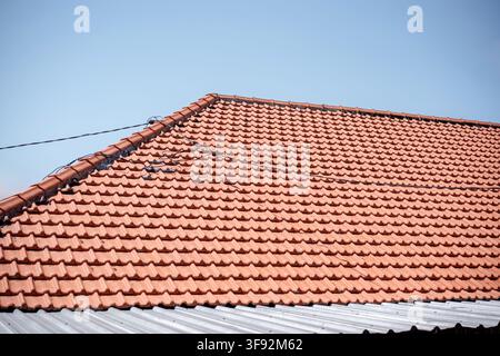 Nahaufnahme von orangefarbenen Terrakottaziegeln auf einem Hausdach vor einem hellen, klaren blauen Himmelshintergrund mit Stromleitungen. Stockfoto