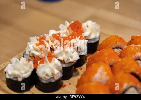 Appetitliche Sushi-Variété, gekrönt mit zarten Sahnespiralen und lebhaften Fischaeiern, präsentiert auf einem natürlichen Holzbrett, das authentische kulinarische Kunst zeigt Stockfoto