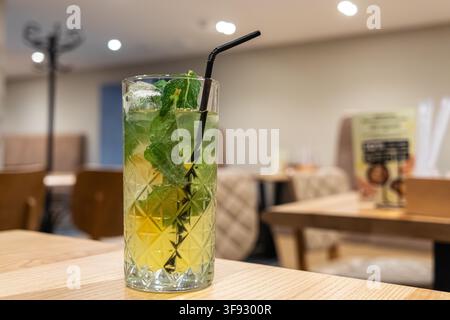 Kaltes Getränk, garniert mit grünen Blättern und Eiswürfeln, serviert in einem Highball-Glas auf einem Café-Tisch, das eine einladende Atmosphäre für Freizeit und geselliges Abendessen schafft Stockfoto