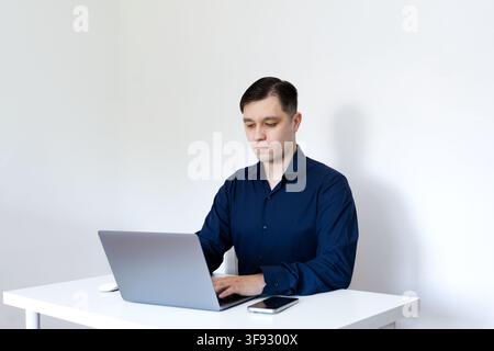Ein junger Berufstätiger trägt ein dunkelblaues Hemd, sitzt an einem sauberen weißen Schreibtisch und konzentriert sich auf die Arbeit an einem grauen Laptop mit einem Smartphone in der Nähe. Stockfoto