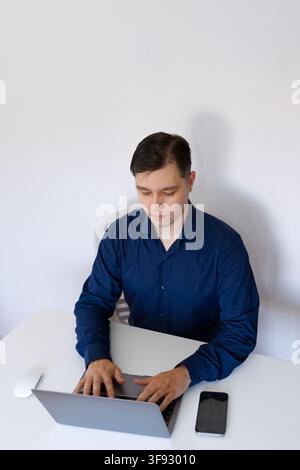 Ein junger Berufsmann trägt ein dunkelblaues Hemd, sitzt an einem sauberen minimalistischen weißen Schreibtisch und konzentriert sich auf die Arbeit an einem grauen Laptop mit einem Smartphone Stockfoto