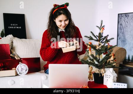 Junge Frau mit Weihnachtsmannmütze hält ein eingewickeltes Geschenk und lächelt auf den Laptop-Bildschirm während eines Videoanrufs zu Hause. Fernbeziehung zu Weihnachten, virtuelles Date Stockfoto
