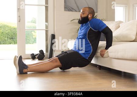 Afroamerikanischer Mann führt Trizeps-Dips auf dem Sofa zu Hause durch, trägt Sportbekleidung mit Hanteln in der Nähe. Homefitness, Gewichte, Training, Athlet, Wohnraum Stockfoto