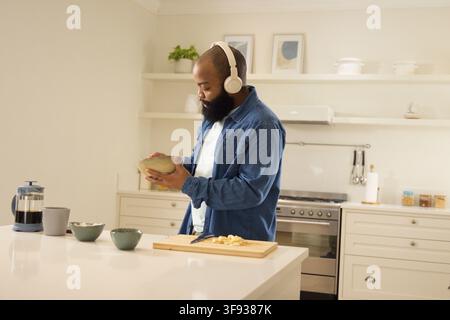 Erwachener afroamerikanischer Mann in Jeans und Headset, der eine Rührschüssel hält und in der Küche Bananen macht Stockfoto