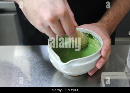 Eine Nahaufnahme einer Person, die einen Bambus-Schnurrer verwendet, um leuchtend grünes Matcha-Pulver in einer Keramikschüssel zu vermischen. Stockfoto