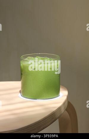 Ein klares Glas Eismatcha-Latte, das auf einem Holztisch in der Nähe eines Fensters steht, beleuchtet von weichem natürlichem Sonnenlicht während des Tages. Stockfoto