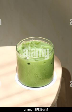 Ein klares Glas Eismatcha-Latte, das auf einem Holztisch in der Nähe eines Fensters steht, beleuchtet von weichem natürlichem Sonnenlicht während des Tages. Stockfoto