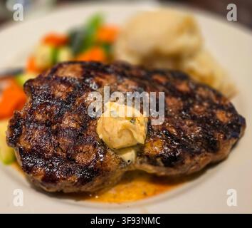 Saftiges gegrilltes Ribeye-Steak mit schmelzender Kräuterbutter, serviert mit cremigen Kartoffelpüree und bunten gebratenen Gemüsen auf einem weißen Teller, e Stockfoto