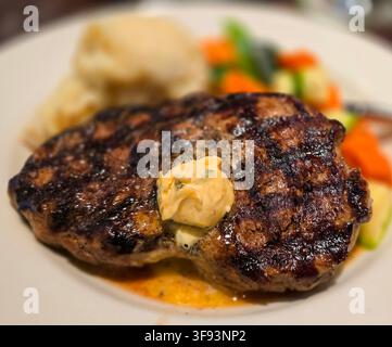 Saftiges gegrilltes Ribeye-Steak mit schmelzender Kräuterbutter, serviert mit cremigen Kartoffelpüree und bunten gebratenen Gemüsen auf einem weißen Teller, e Stockfoto
