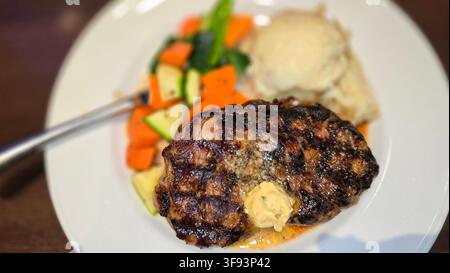 Saftiges gegrilltes Ribeye-Steak mit schmelzender Kräuterbutter, serviert mit cremigen Kartoffelpüree und bunten gebratenen Gemüsen auf einem weißen Teller, e Stockfoto