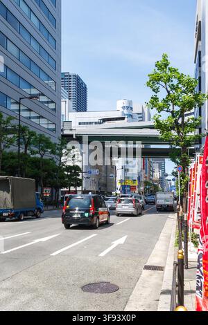 Belebte städtische Straßenansicht mit Autos, die unter einer erhöhten Autobahnbrücke zwischen modernen Wolkenkratzern in der Innenstadt von Osaka, Japan, fahren. Stockfoto