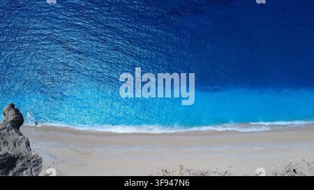 Atemberaubender Luft-Drohnenaufnahm von kristallklaren türkisfarbenen Meereswellen, die auf einen weißen Sandstrand mit felsigen Klippen treffen. Stockfoto