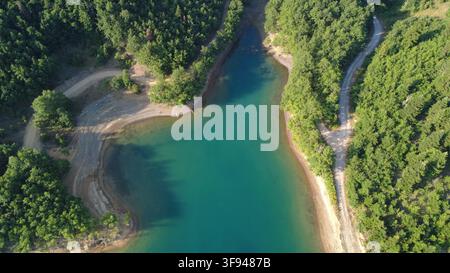 Luftbilddrohnenaufnahme einer atemberaubenden türkisfarbenen Lagune, umgeben von dichtem grünem Wald, mit sich windenden Schotterwegen, die zum Ufer führen. Stockfoto
