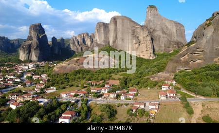 Luftaufnahme der dramatischen Meteora-Felsformationen, die über einem traditionellen griechischen Dorf thronen, das sich zwischen üppigen grünen Hügeln unter einem strahlend blauen Himmel befindet. Stockfoto