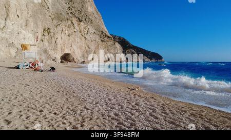 Dramatische Kalksteinklippen ragen über einen abgelegenen Kiesstrand mit türkisfarbenen Wellen, die unter einem klaren blauen Himmel an das Ufer schlagen. Ein klassisches Mittelmeer Stockfoto