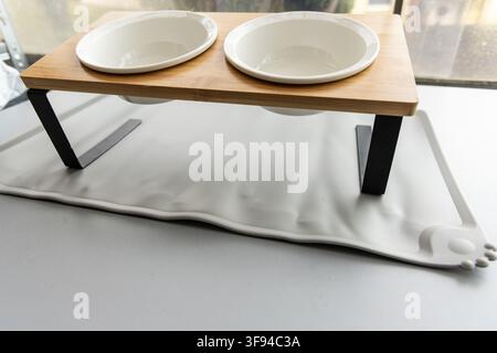 Ein erhöhter doppelter Haustierfutterspender mit weißen Keramikschüsseln steht auf einer grauen Matte Stockfoto