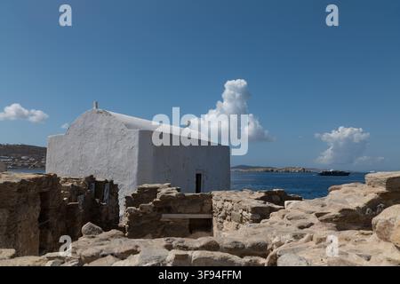 Kleine weiß getünchte Kapelle, die über alten Steinruinen mit dem Meer im Hintergrund steht, Mykonos, Griechenland. Stockfoto