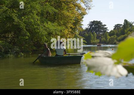 Zwei Personen rudern ein kleines Holzboot auf einem ruhigen See, umgeben von üppigen grünen Bäumen an einem sonnigen Tag. Stockfoto