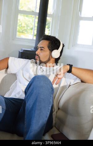 Mittelalter Afrikaner lehnt sich zu Hause auf dem Sofa zurück und trägt weiße Kopfhörer, die eine schwarze Smartwatch zeigen Stockfoto