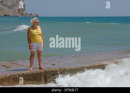 Senior ältere Dame steht auf der Strand-Leiste auf Hintergrund der Wellen planschen. Stockfoto