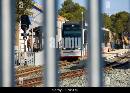 Calp, Spanien - 11. Juli 2027: Blick auf eine moderne Straßenbahn, die am Bahnsteigdamm ankommt, mit Fahrgästen, die unter einem Unterstand warten, gesehen durch einen Metallzaun Stockfoto