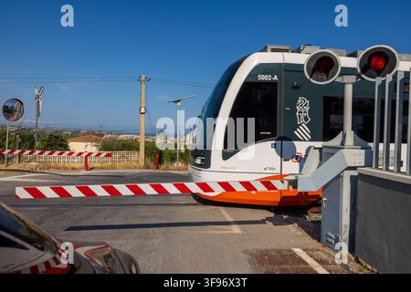 Calp, Spanien - 11. Juli 2027: Blick auf eine moderne Straßenbahn, die eine Eisenbahnüberführung mit roten und weißen Schranken passiert, klarer blauer Himmel und Küstenlandschaft Stockfoto