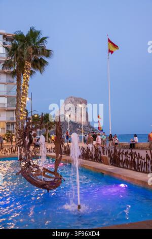 Calp, Spanien - 11. Juli 2027: Blick auf den Penon de Ifach, der über der Küstenpromenade aufragt, mit einem dekorativen Brunnen, Palmen und Menschen, die die Stockfoto