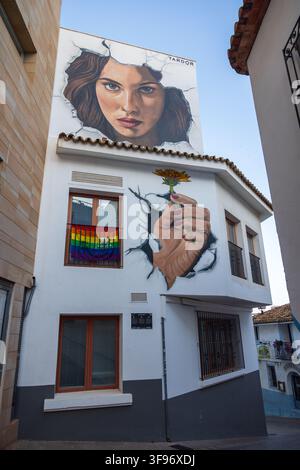 Calp, Spanien - 11. Juli 2027: Blick auf ein großes Wandgemälde mit dem Gesicht einer Frau und einer Hand, die eine Blume hält, das auf der weißen Fassade eines Gebäudes in th Stockfoto