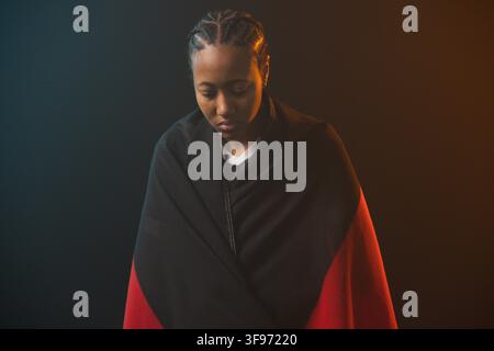 Afroamerikanische Frau posiert im Studio, trägt schwarzen roten Schal, blickt unter bernsteinfarbenem Türkislicht nach unten Stockfoto