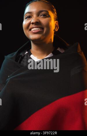 Lächelnde afroamerikanische Frau in einem Trikot, eingewickelt in eine schwarz-rote Decke, Studio-beleuchteter Teal-Ambersaum Stockfoto