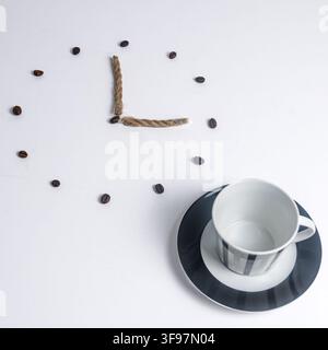 Oberansicht einer leeren gestreiften Tasse und Untertasse neben einer minimalistischen Uhr aus Jutegarn und verstreuten gerösteten Kaffeebohnen auf weißem Grund. Stockfoto