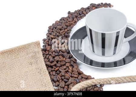 Nahaufnahme eines leeren gestreiften Kaffeebechers und Untertassen neben gerösteten Bohnen, Sackleinen und Jute-Seil auf einem hellen weißen Hintergrund. Stockfoto