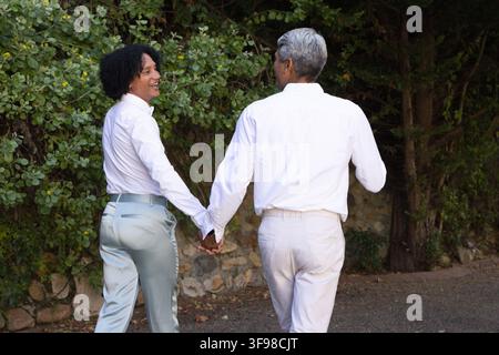 Afrikanische Männer, die sich an den Händen halten und einen Kiesweg entlang einer niedrigen Steinmauer entlanggehen, tragen weiße Hemden, Gürtel Stockfoto