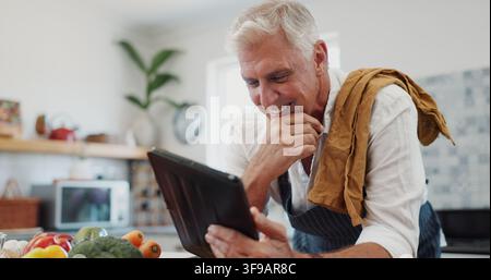 Senior, Mann und Kochen in der Küche mit Tablet, lächelt und durchsucht das Internet, um online ein Rezept zu recherchieren. Glücklicher, älterer Mensch und Technik zu Hause mit Stockfoto