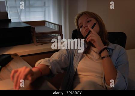 Junge Frau sitzt am Schreibtisch, sieht unmotiviert und müde aus, während sie einen Stift am Kinn hält, kämpft mit Konzentration und Prokrastination während Stockfoto