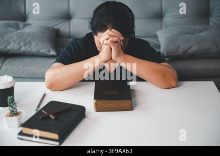 Mittealterne christliche Frau betet mit gesenktem Kopf und gefalteten Händen an einem Tisch mit einer Heiligen Bibel, Büchern und einem Holzkreuz und drückt Glauben, Hoffnung, s aus Stockfoto