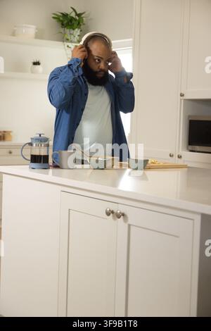 Afroamerikanischer Mann in Jeanshemd, der zu Hause Kopfhörer anpasst und Kaffee, French Press, Toast macht Stockfoto