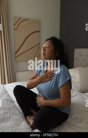 Seniorasiatische Frau in blauem Oberteil und schwarzen Leggings sitzt auf dem Bett und übt Atemübungen mit Bettdecke Stockfoto