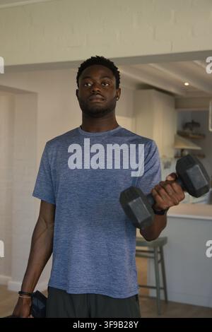 Afrikanischer Mann hebt Sechskant-Hanteln, trägt blaues T-Shirt und Shorts in einer offenen Küche mit Armband Stockfoto