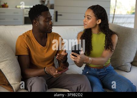 Afroamerikanisches Paar sitzt zu Hause auf dem Sofa, in orangefarbenem T-Shirt und Limettengrünem Oberteil, zeigt Handys Stockfoto