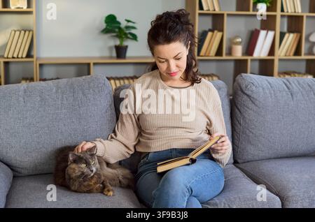 Ruhige Frau liest ein Buch neben ihrer Katze auf dem Sofa zu Hause, entspannte gemütliche Stimmung Stockfoto