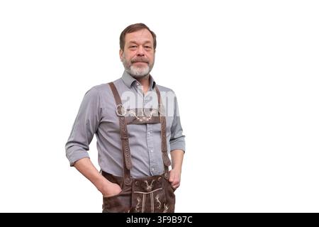 Ein reifer bärtiger Mann in traditioneller Lederhose und einem grauen Hemd steht mit den Händen in den Taschen auf einem hellen Hintergrund. Stockfoto
