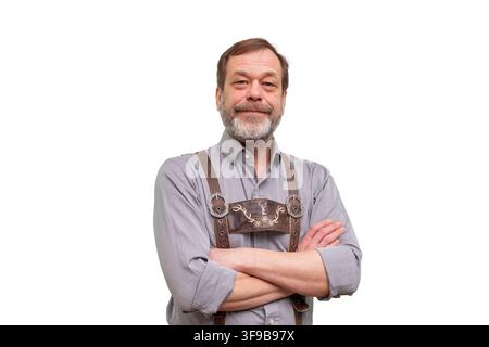 Ein lächelnder, reifer Mann, der traditionelle bayerische Hosenträger trägt, steht mit verschränkten Armen auf einem weißen isolierten Hintergrund. Stockfoto