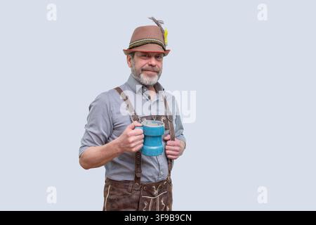 Ein lächelnder bärtiger Mann trägt einen Hut, ein Hemd und eine Lederhose im bayerischen Stil und hält einen blauen Bierkrug vor einem schlichten Hintergrund. Stockfoto