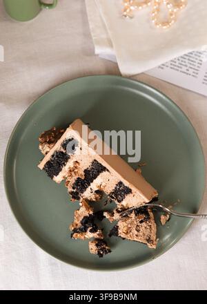 Nahaufnahme einer Scheibe geschichteten Schokoladenkuchens mit cremiger Füllung, serviert auf einem grünen Teller mit einer Gabel und einer Tasse Kaffee im Hintergrund. Stockfoto