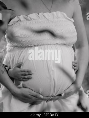 Schwarz-Weiß-Nahaufnahme-Mutterschaftsporträt einer schwangeren Frau, die ihren Babybauch sanft mit beiden Händen im Freien bei natürlichem Licht hält. Stockfoto